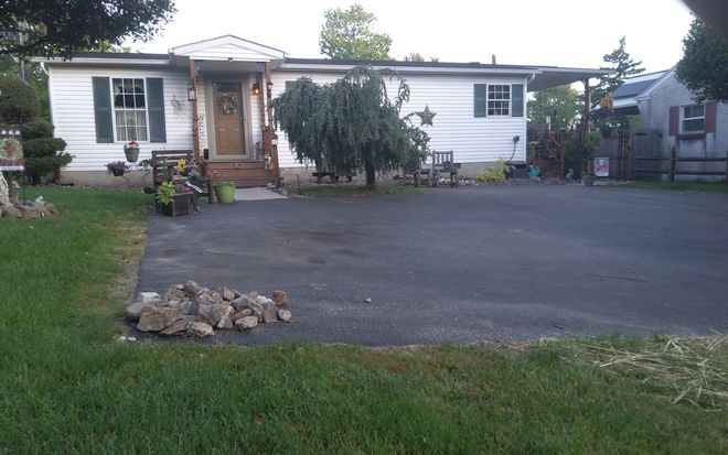 The before image of a single-story white house with a large dark driveway, front steps, and a grassy yard.