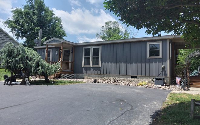 The after image of the gray single-story house with a large driveway, front windows, and trees under a cloudy sky