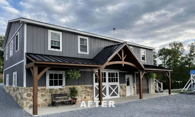 A grey, two-story barn-style building with a stone foundation, black metal roof, and wooden porch with a curved truss.