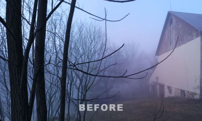 A weathered barn stands in a misty, fog-covered field at dusk, with bare tree branches in the foreground.