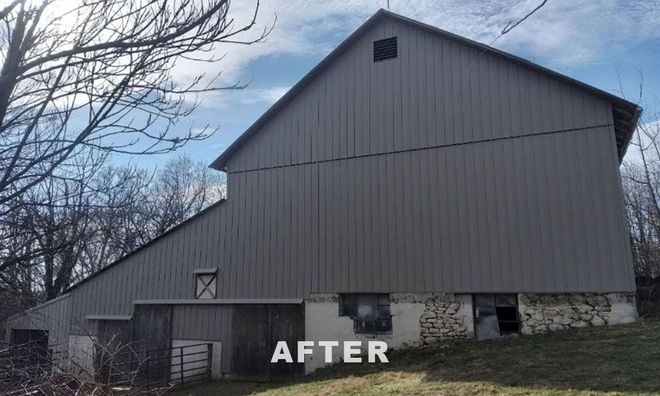 A large barn with grey vertical metal siding and a stone foundation, set against a backdrop of trees under a blue sky.