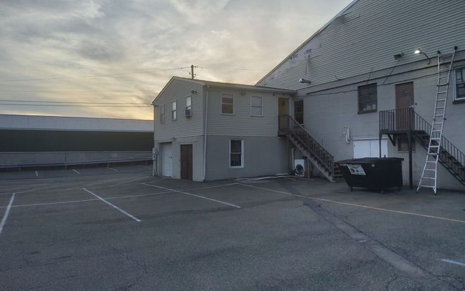 Empty parking lot beside a metal building at sunset, with a dumpster and ladder near the entrance.