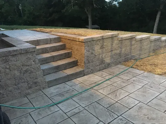 Stone steps and retaining wall in a backyard with a paved patio. Green hose is visible.