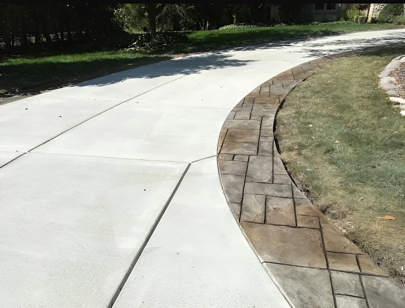 Curving concrete driveway with patterned brick border next to grassy lawn.