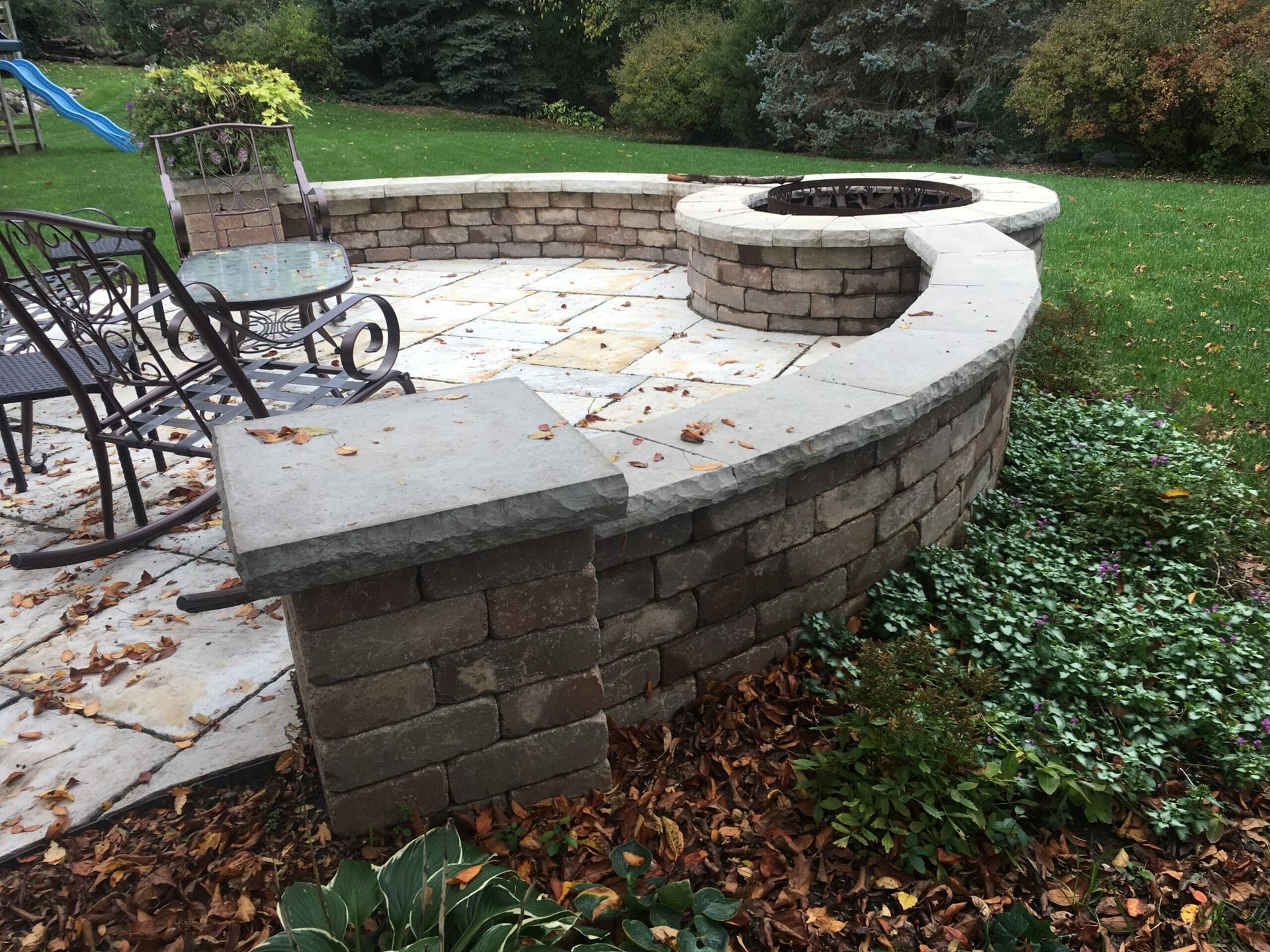 Stone patio with fire pit, seating area, and landscaping in a backyard.