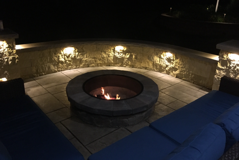Fire pit with flames, surrounded by stone wall with lights and blue lounge seating at night.