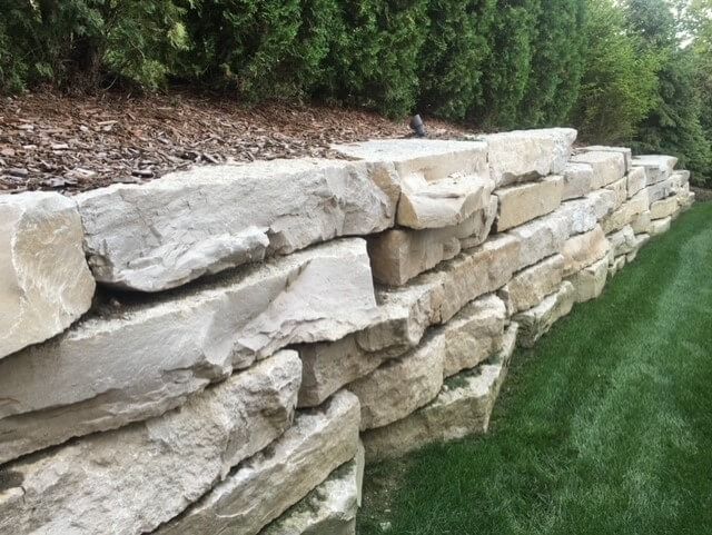 Stone retaining wall along a grassy lawn, with trees and mulch in the background.