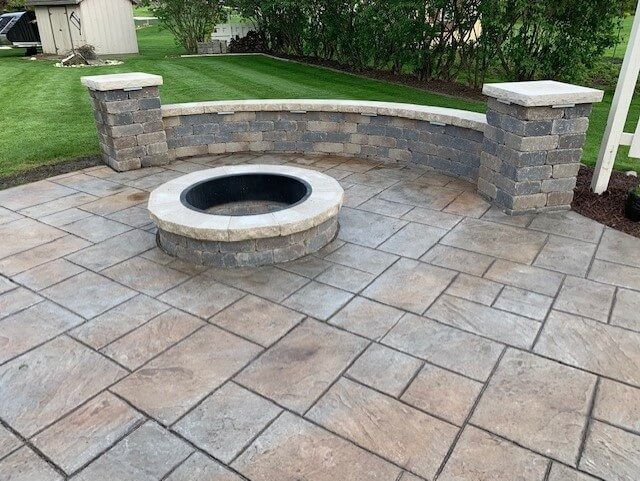 Stone patio with fire pit, curved seating wall, and pillars, in a backyard setting.