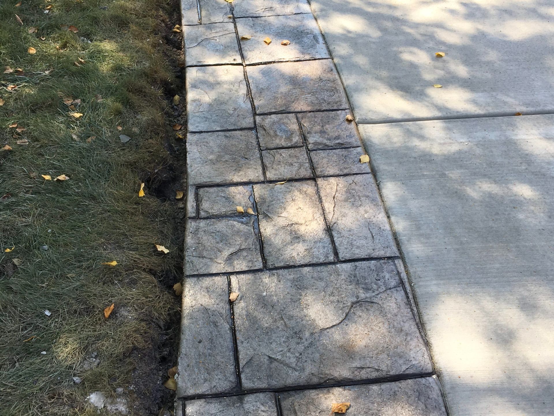 Concrete walkway next to grass and a driveway. The walkway has a textured stone pattern.