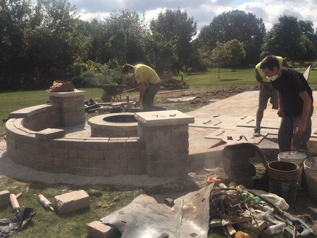 Construction workers building a brick fire pit and patio outdoors on a sunny day.