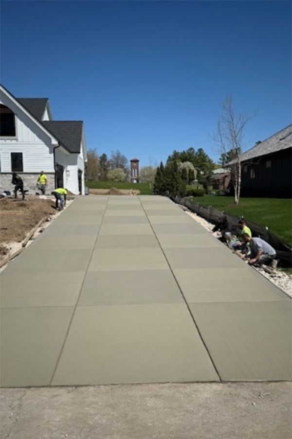 Newly poured concrete driveway with workers, house on the left, and a tower in the distance under a blue sky.