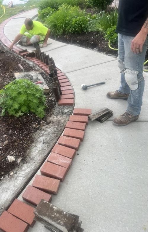 Two workers installing a brick border along a concrete walkway near landscaping.