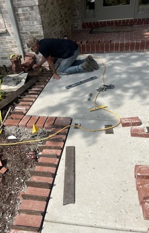 Person installing brick border around a concrete walkway. Bricks are red. Tools and materials are present.