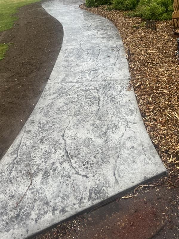 Stamped concrete walkway curves through a yard; gray stone texture, dark soil, and wood chips alongside.