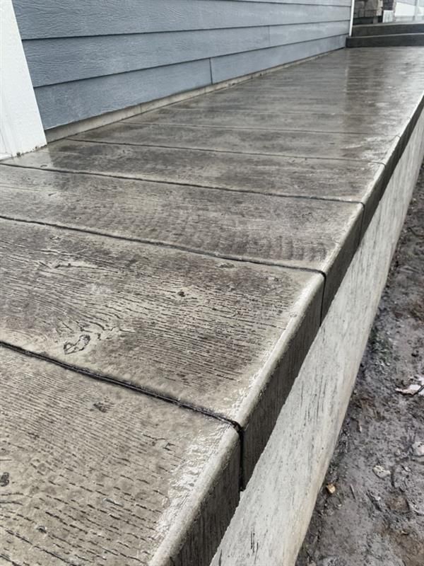 Concrete walkway with textured wooden plank design next to a house with blue siding.