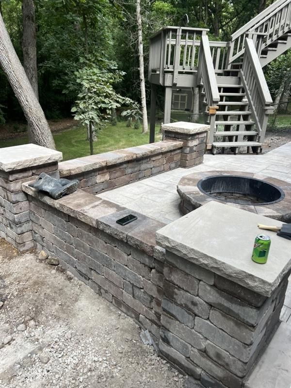 Stone patio with built-in fire pit, low walls, and wooden stairs leading to a structure in a wooded area.