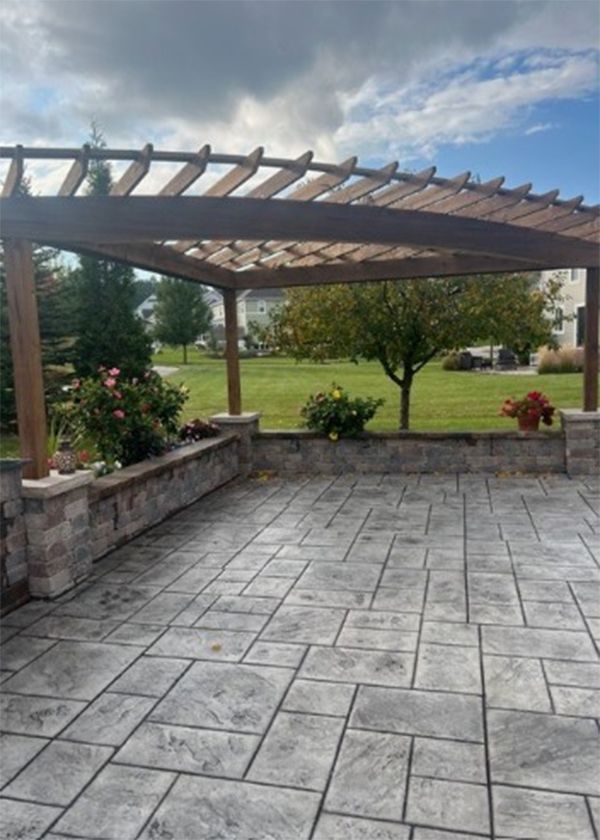 Patio with pergola, stone wall, and patterned gray floor. Green grass and trees in the background.
