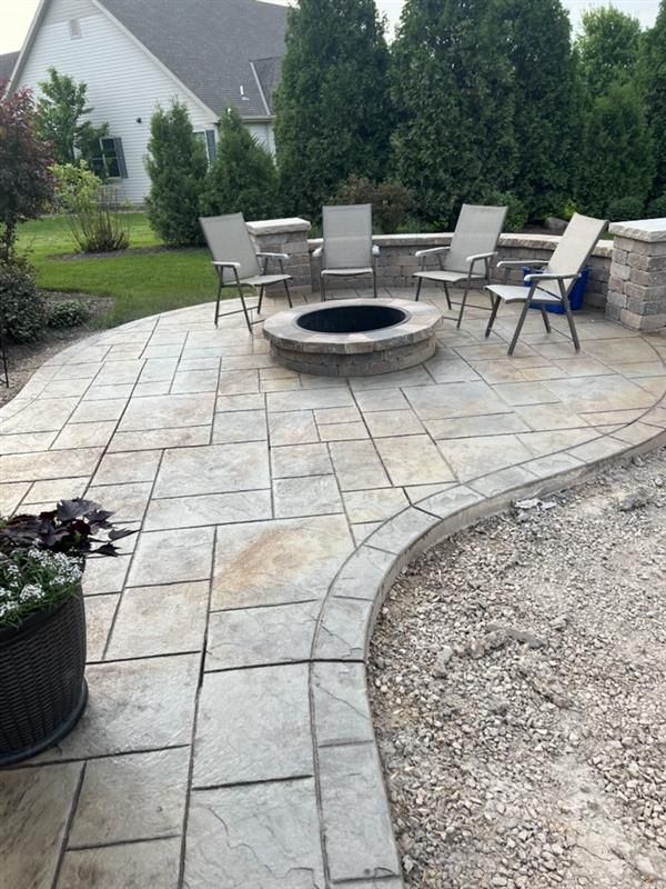 Outdoor patio with fire pit and seating, stone pavers, surrounding greenery, and a house in the background.