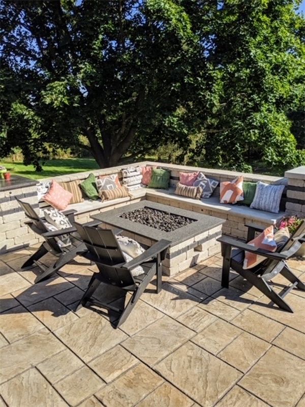 Outdoor patio with a stone fire pit, seating, and decorative pillows under a large tree.