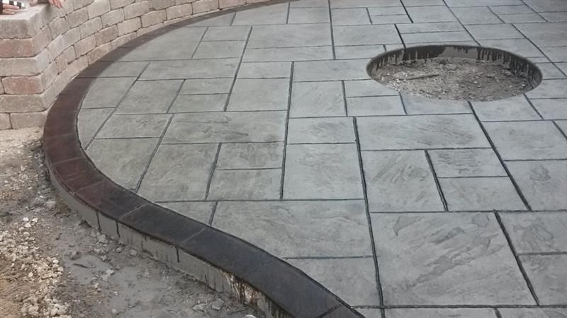 Curved gray patio with fire pit, bordered by dark edging and a stone wall.