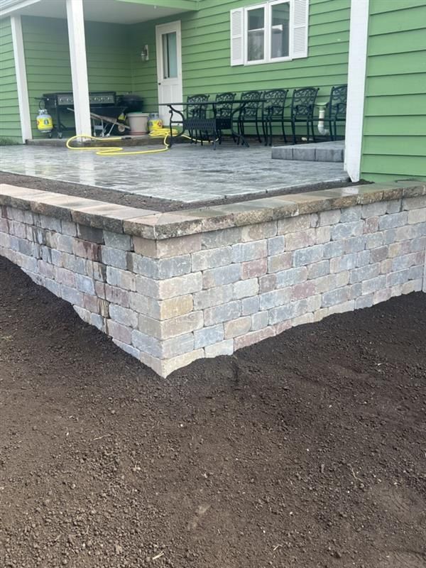 Brick retaining wall bordering a paved patio. Green house in background.