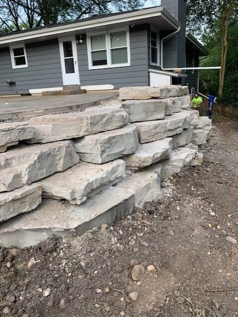 Stone retaining wall in front of a gray house. Dirt ground and two workers nearby.