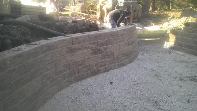 A curved retaining wall made of brown bricks is being built. A worker is seen on the wall.