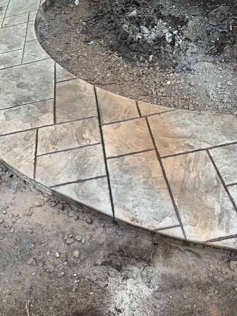 Concrete patio with textured, light-colored tiles in a curved design, partially surrounded by dirt.