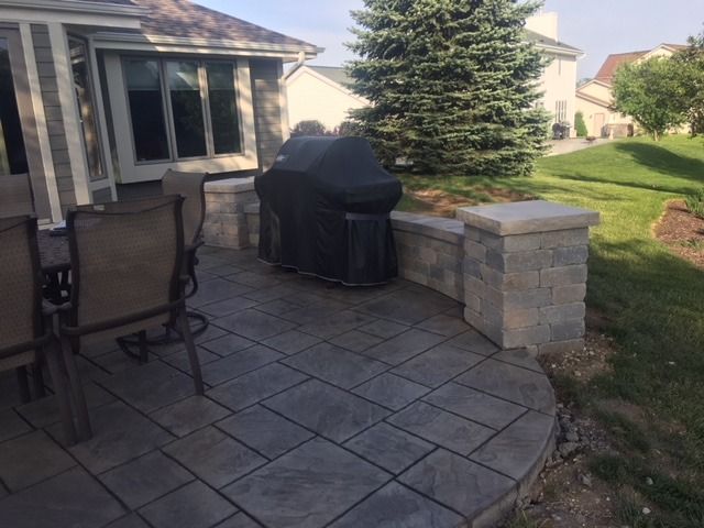 Stone patio with a grill covered by a black cover, seating, and a stone retaining wall.