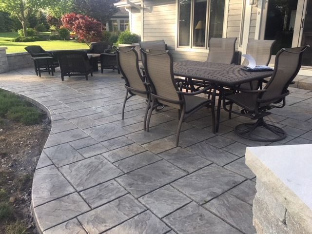 Outdoor patio with a stone-like surface and a dining table with chairs. Additional seating in the background.