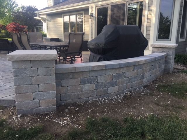 Stone retaining wall with grill, patio furniture, and house in the background.