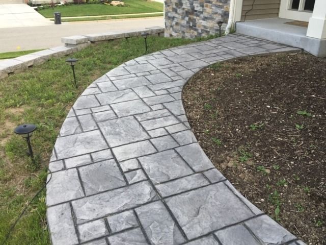 Curving gray stone pathway leading to a house entrance. Green grass and garden beds on either side.