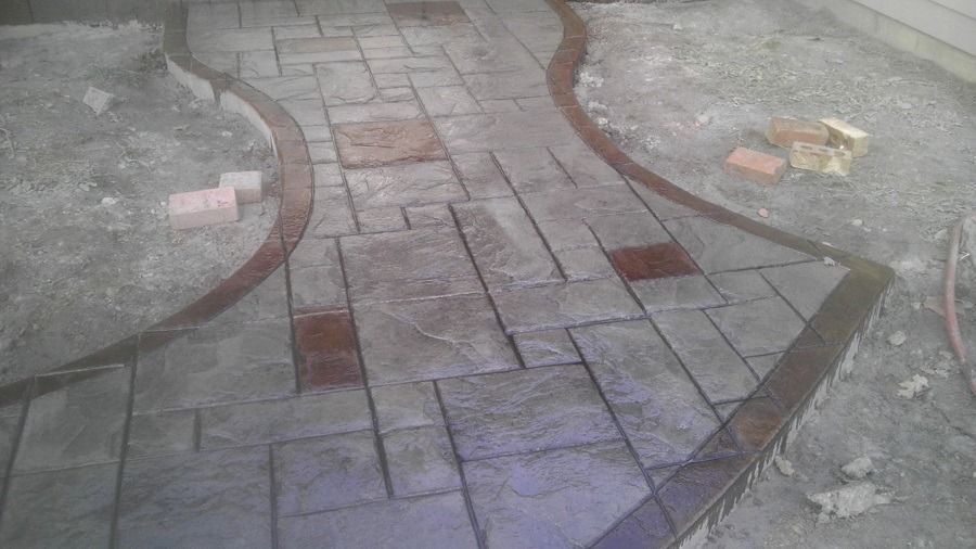 Concrete pathway with a dark brown border and brick accents, in a construction setting.