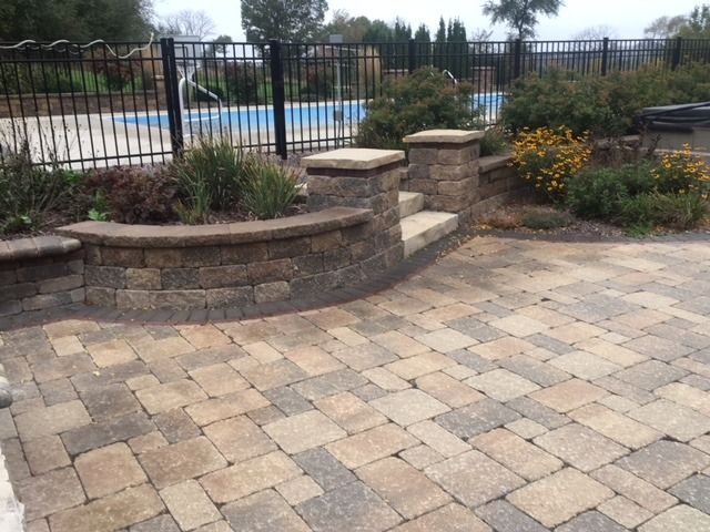 Brick patio with steps, flowerbeds, and pool behind a black fence.