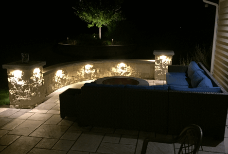 Outdoor patio at night, illuminated stone wall with embedded lights, sofa, fire pit, tree in background.