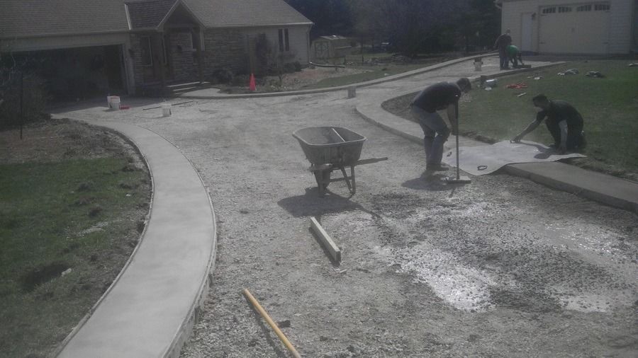 Construction workers leveling concrete for a driveway. Wheelbarrow and house visible.
