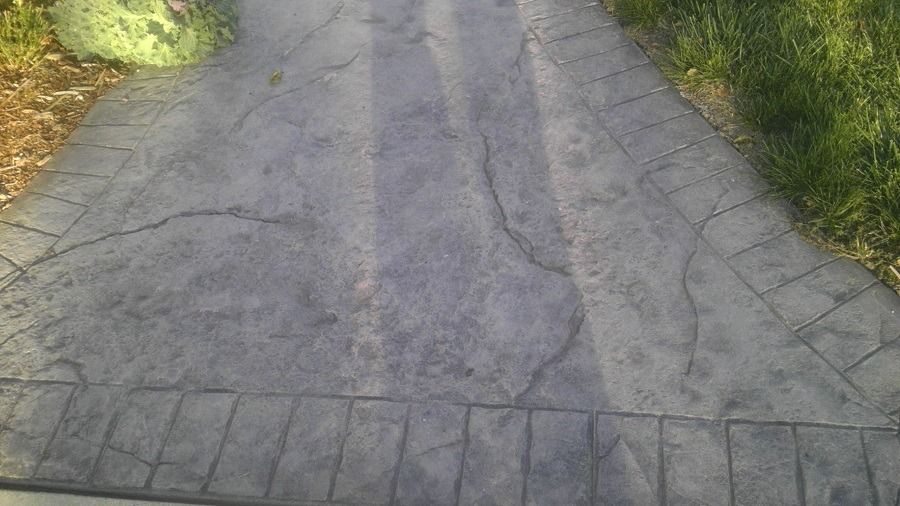 Gray, textured concrete driveway with a brick border, cracks, and grass on the right.