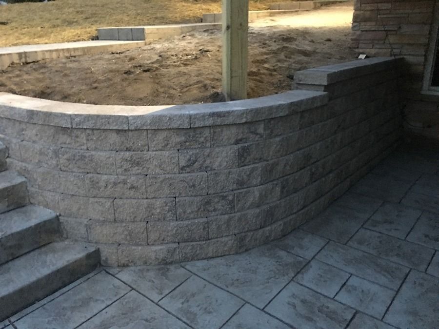 Curved retaining wall made of gray blocks, next to steps and a patio with textured concrete.