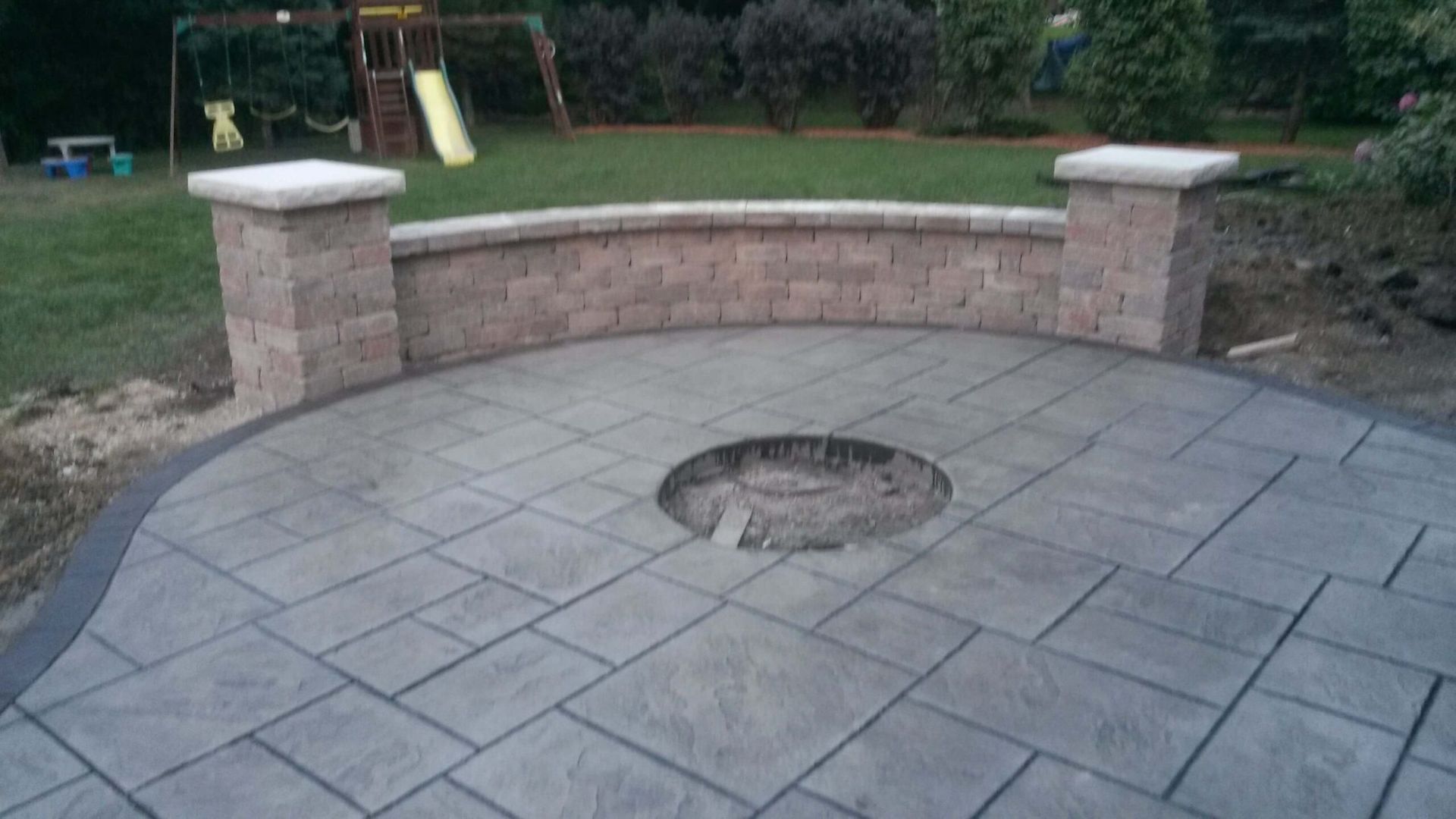 Stone patio with built-in fire pit, surrounded by a low brick wall and columns, set in a backyard.