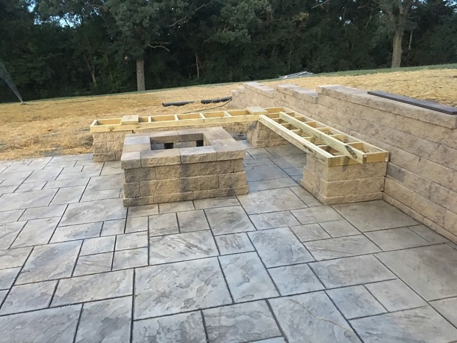 Stone patio with a built-in fire pit and seating under construction in a yard.