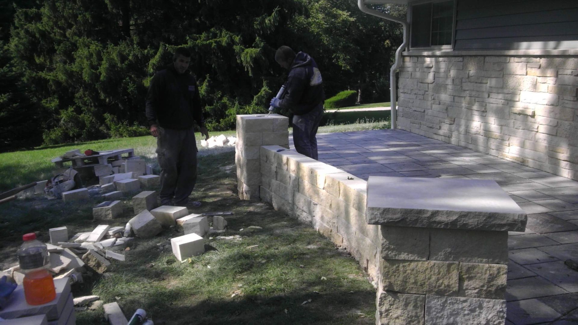 Two workers building a stone wall on a patio. Green lawn, trees in background.
