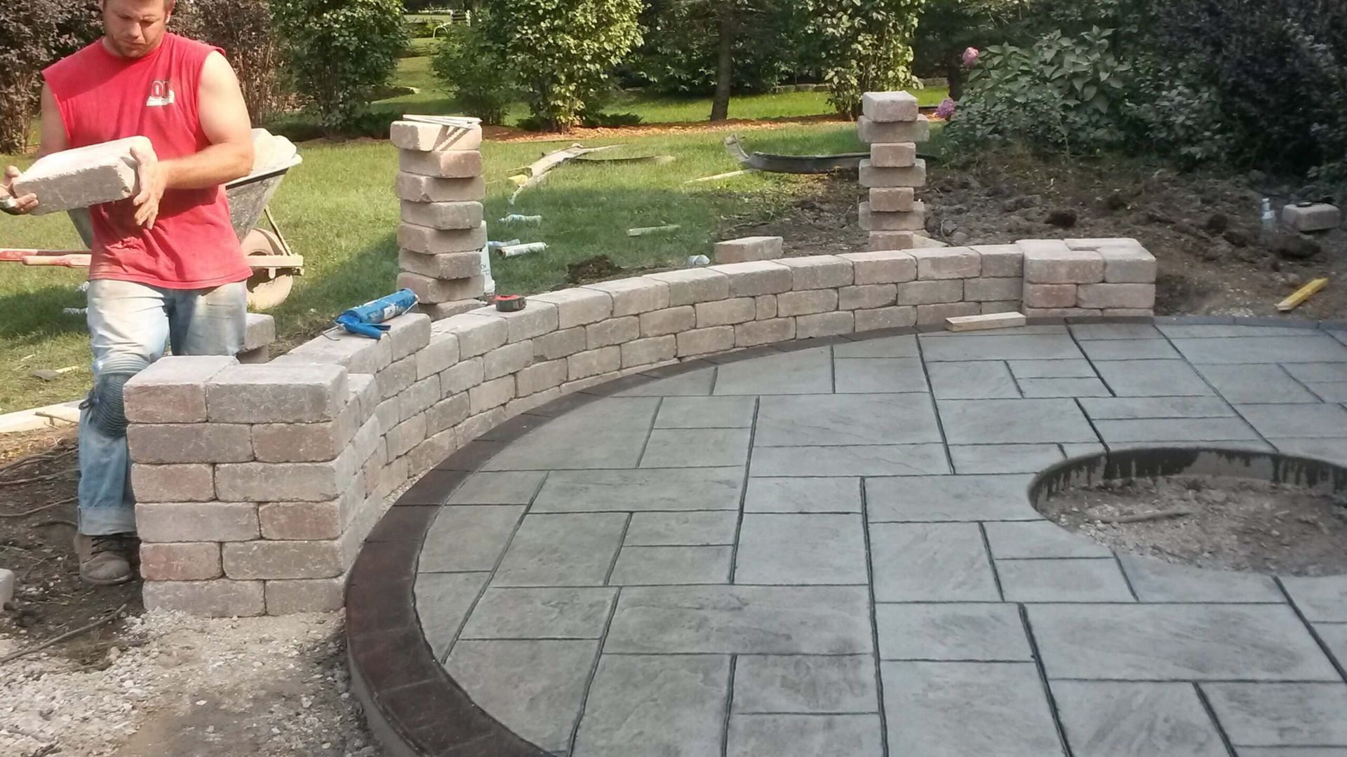 Man building a brick retaining wall around a patio with fire pit.