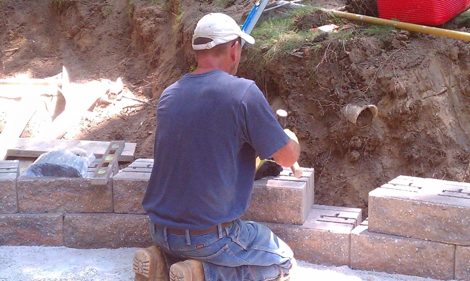 Man kneeling, building a retaining wall with brown blocks, near dirt bank and pipe.