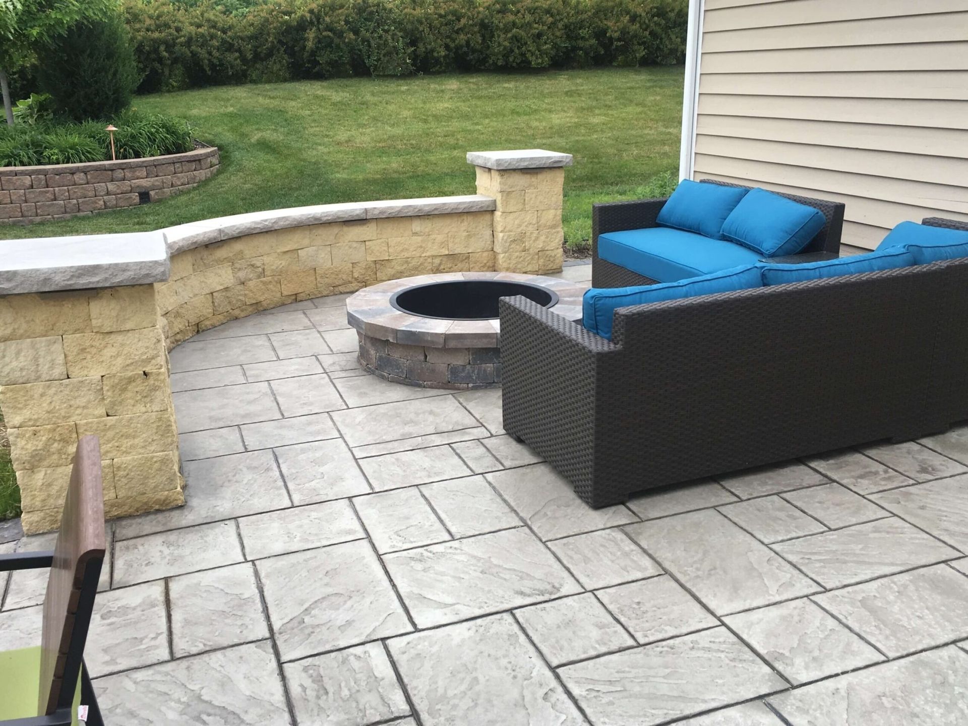 Outdoor patio with seating, fire pit, and retaining wall. Gray pavers, blue cushions, green lawn.