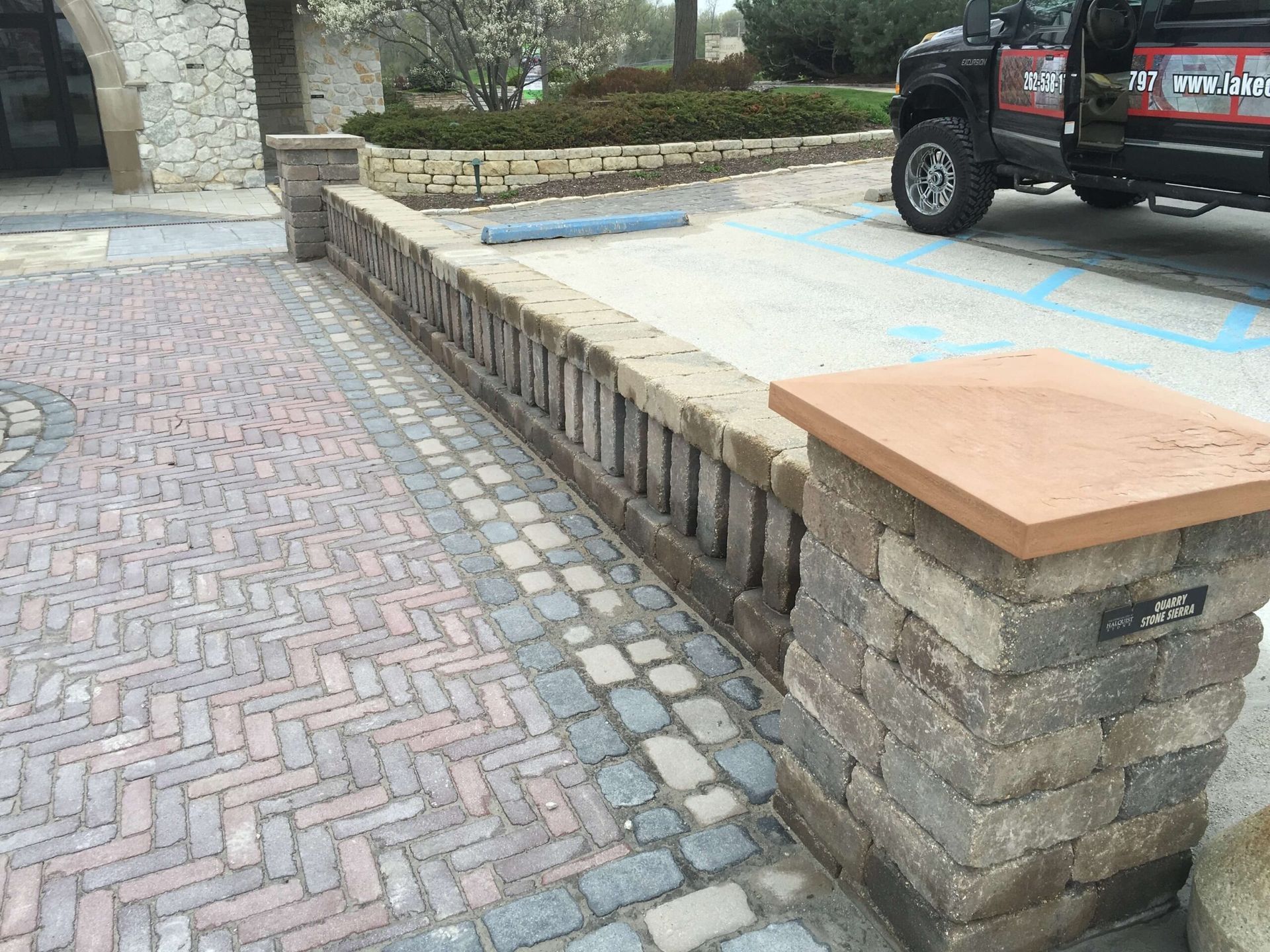 Brick wall and pillar with a stone top and a brick paved area next to a blue parking space.