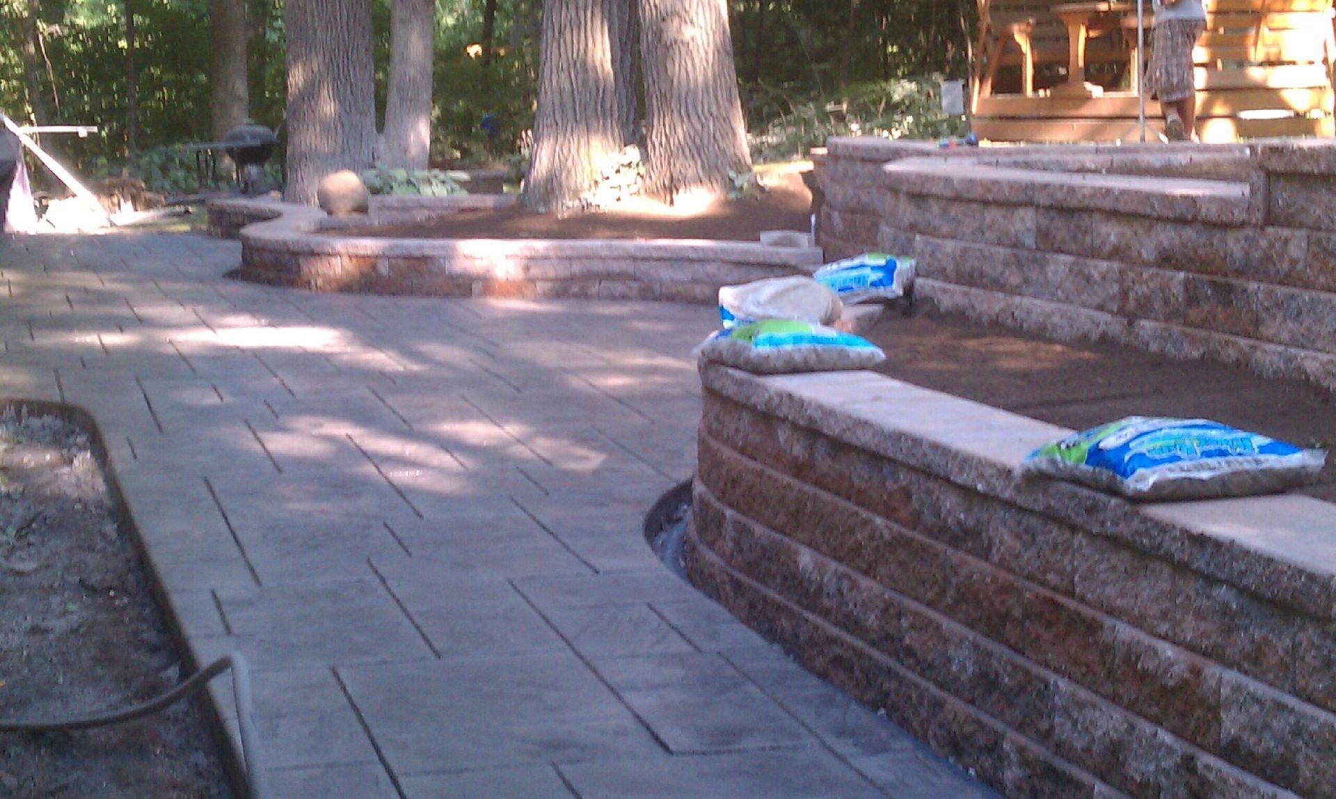 Stone patio with tiered retaining wall. Blue bags sit on the wall. Trees in the background.