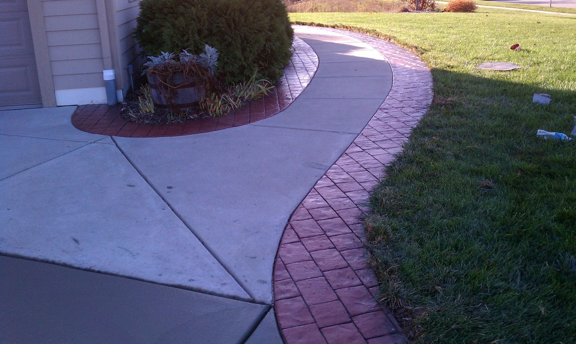 Concrete driveway and walkway with brick-pattern border, next to green grass and a bush.