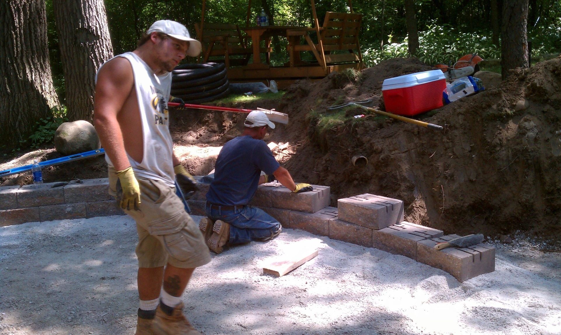 Two men constructing a brick wall on a bed of gravel outdoors, with a red cooler nearby.
