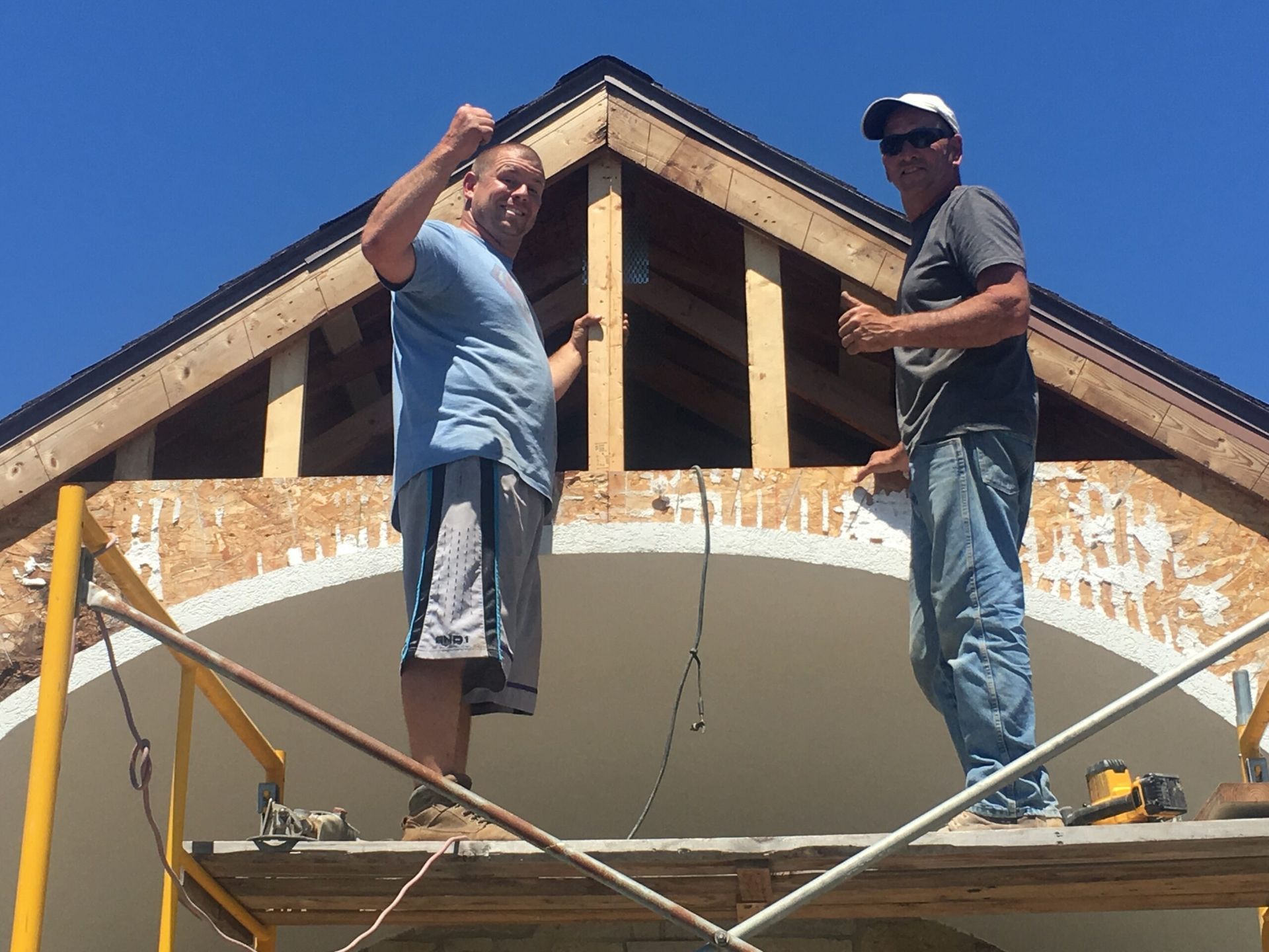 Two men on scaffolding, pose with arms raised in front of a gable roof frame. Bright sunny day.