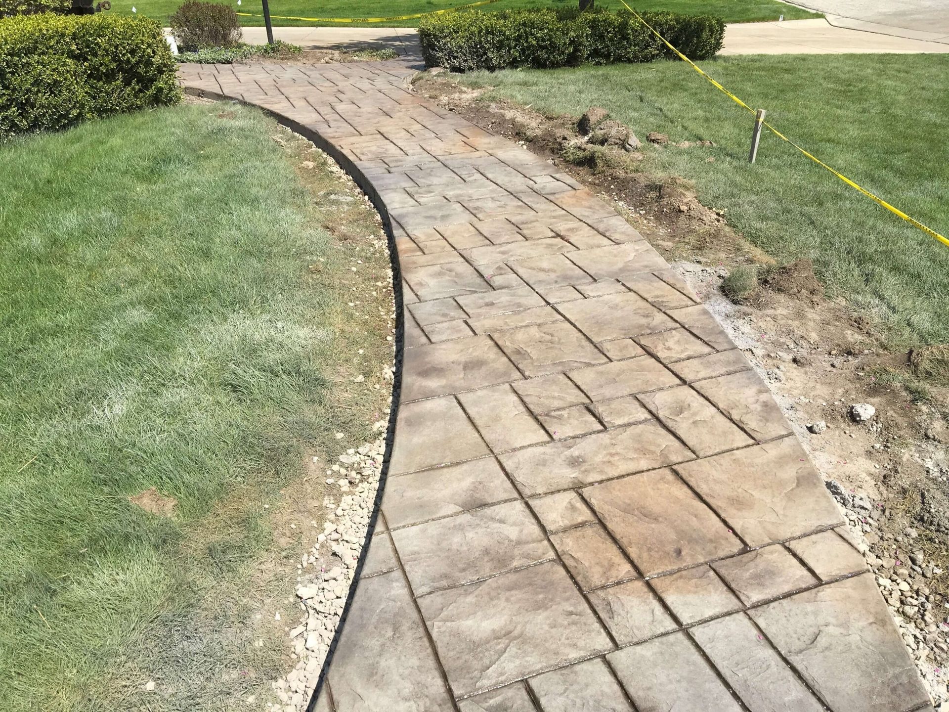 Stone paver walkway curves through a grassy yard, with a gravel edge and a yellow construction tape in the background.